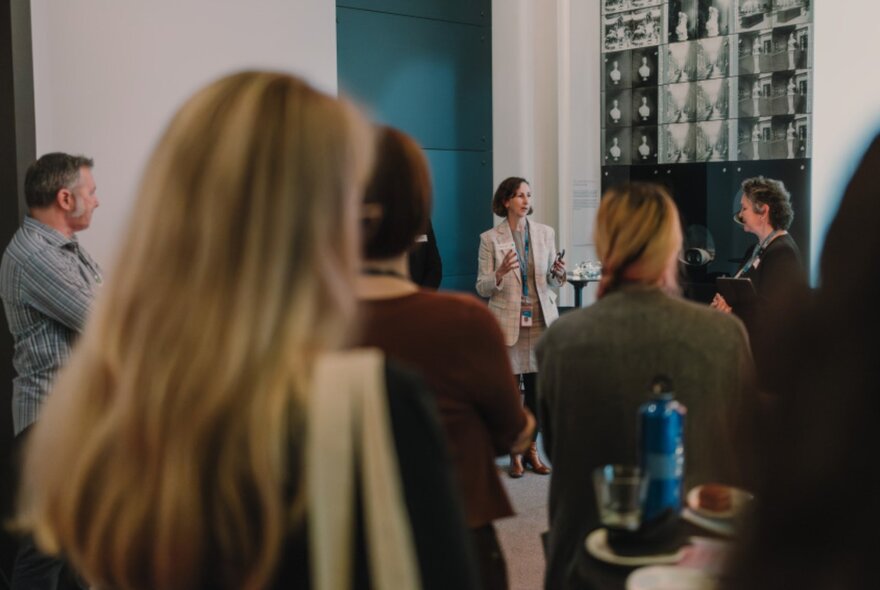 People casually standing in a room looking a woman in a jacket who is presenting. 