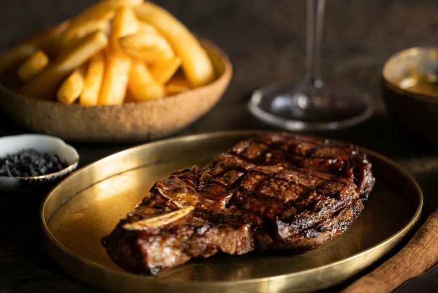 Hearty steak on golden plate, with dish of fries at rear.