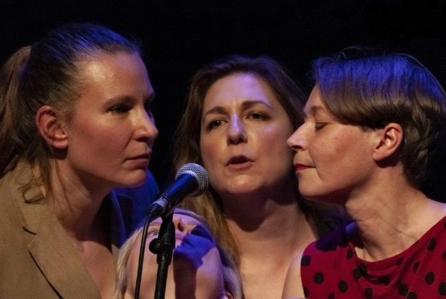 Three female improvisers on stage, with their heads close together, leaning into a microphone.