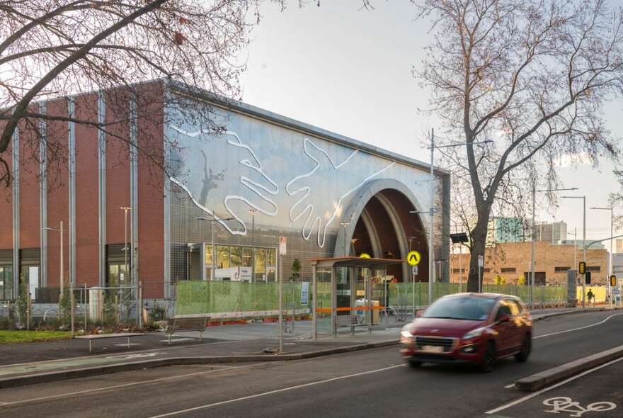 Artwork on the exterior facade of a square red brick building with a large archway, depicting two hands coming together; a red car driving on the road in front.