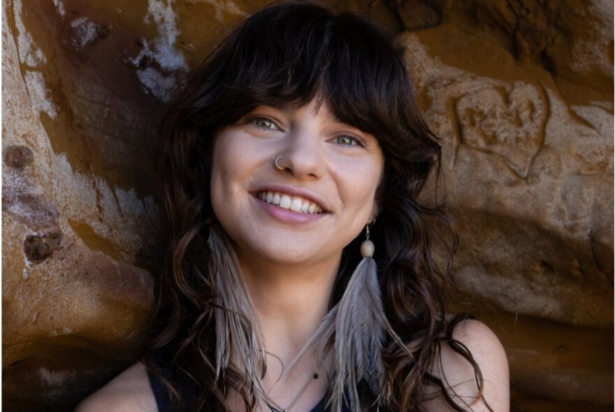 A smiling woman with dark brown hair and a heavy fringe, wearing long, light-grey fringe earrings. standing outdoors in front of a textured stone background.