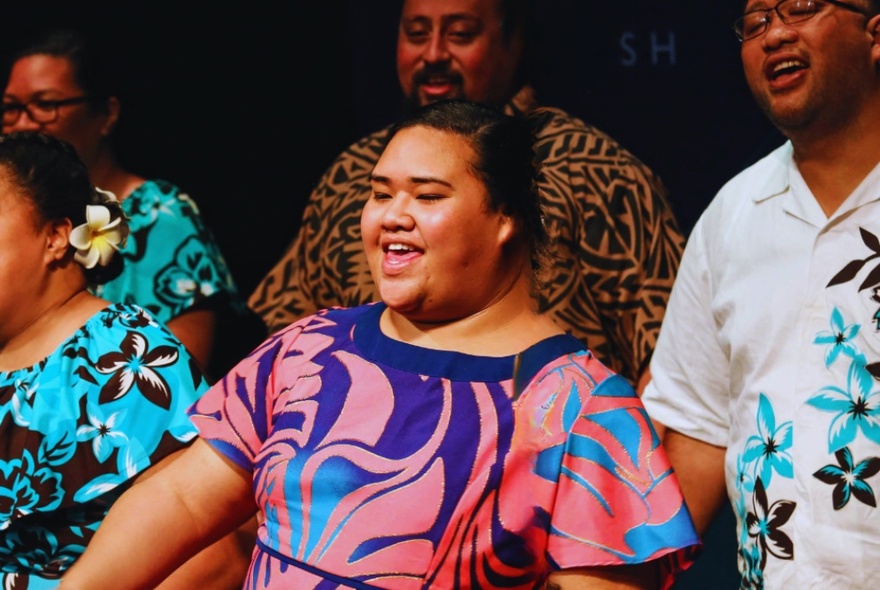 A group of Pacific Islander people in patterned dresses and shirts singing on stage. 