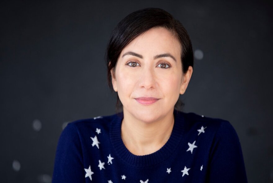 Writer Sarah Krasnostein, her dark hair pulled away from her face, smiling slightly an wearing a navy blue top with a white star pattern, seated in front of a dark grey backdrop.