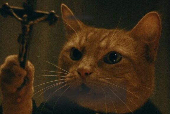 A ginger cat wearing a black cape and holding up a crucifix in his ginger paw.