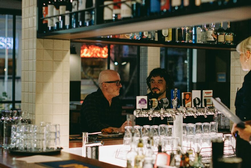 Two friends seated and enjoying a drink at a bar, with bottles, glasses and the bar service area in the foreground.