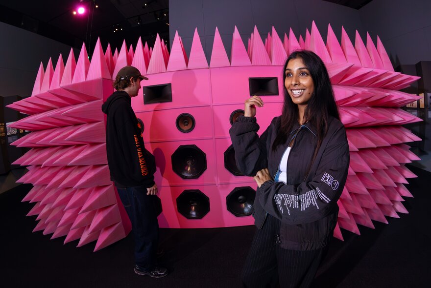 Two people standing in front of a large pink spiky wall of speakers in an exhibition space.