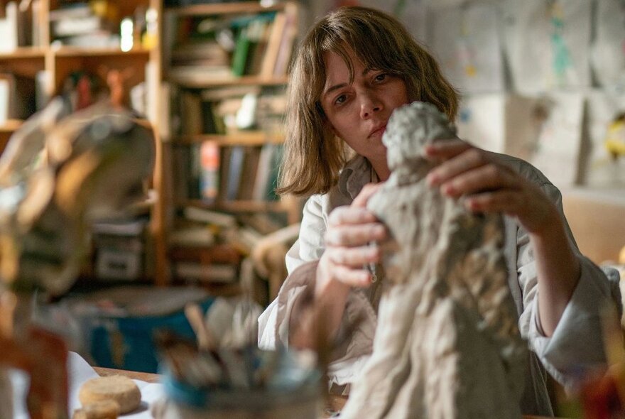 A scene from a film showing a woman creating a sculpture in a workshop, a messy bookshelf behind her. 