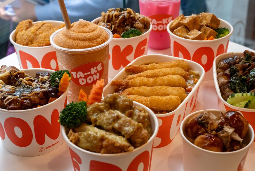 An assortment of Japanese rice bowls in takeaway containers, a side of takoyaki and two cold drinks.