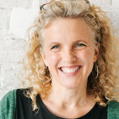 Children's author Sally Rippin, smiling with curly blonde hair, against a wall of cream bricks.