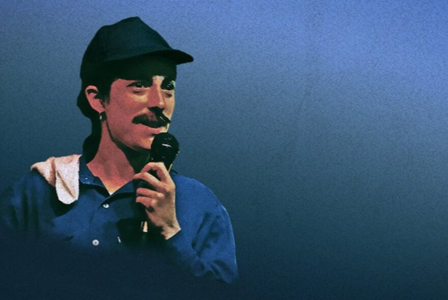 Performer wearing a cap, and a blue boiler suit, with a thick moustache, talking into a microphone, against a blue background.