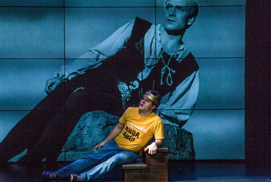 A performer on stage, reclining on a set of wooden stairs, with an image of a person in a similar pose projected onto a screen behind him.