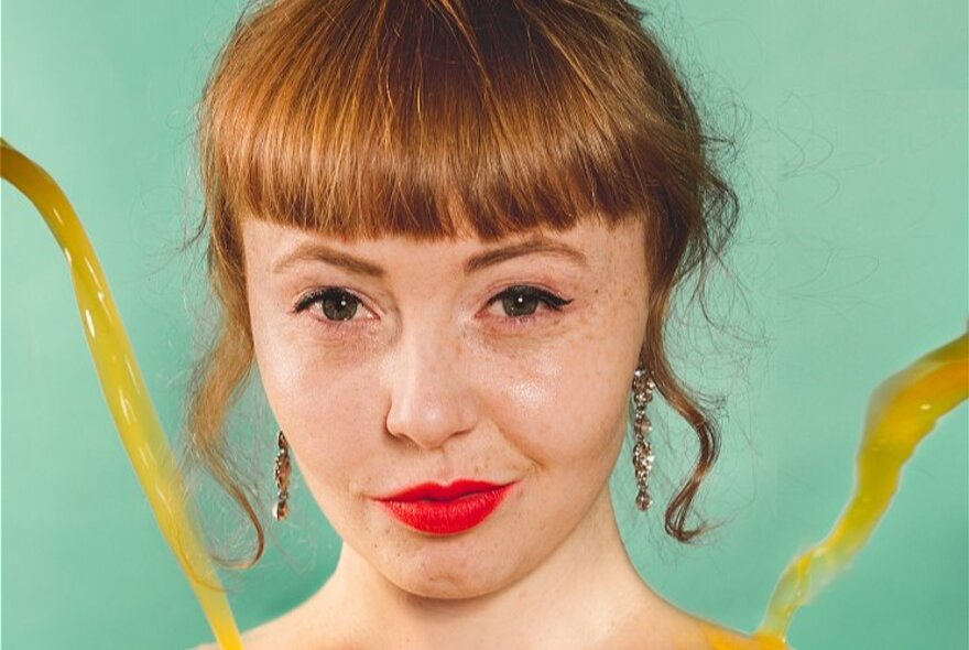 A woman with a blunt ginger coloured fringe, wearing red lipstick and posed against a pale green background, two streams of orange juice in the foreground framing her face.