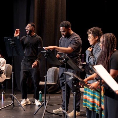 Five actors seated in chairs on a stage at a live play reading. 