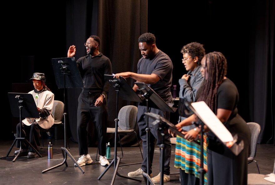 Five actors seated in chairs on a stage at a live play reading. 