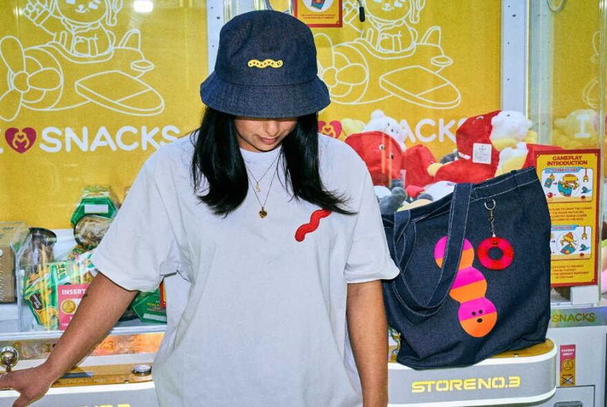 A person wearing a white t-shirt and blue bucket hat standing at a merchandise stall at Melbourne Central, with objects on display such as a tote bag and caps.