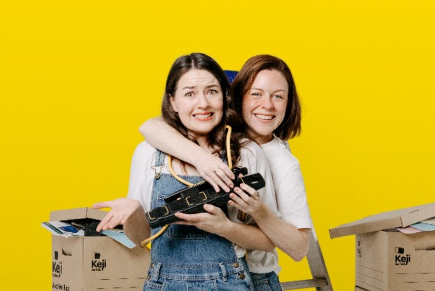 Two women, one with her arm over the shoulder of the other as they hold up a piece of electrical equipment with moving boxes behind them. 