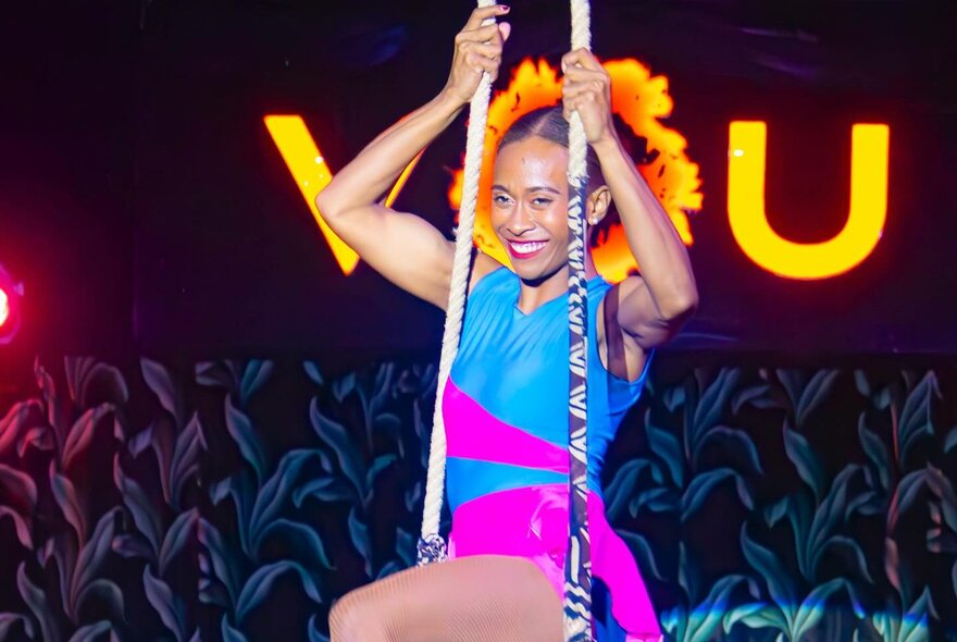 A circus performer on a trapeze, smiling, in front of a VOU sign. 