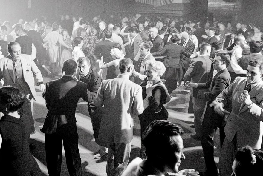 An old black and white image of people at a large dance hall all paired up and dancing.
