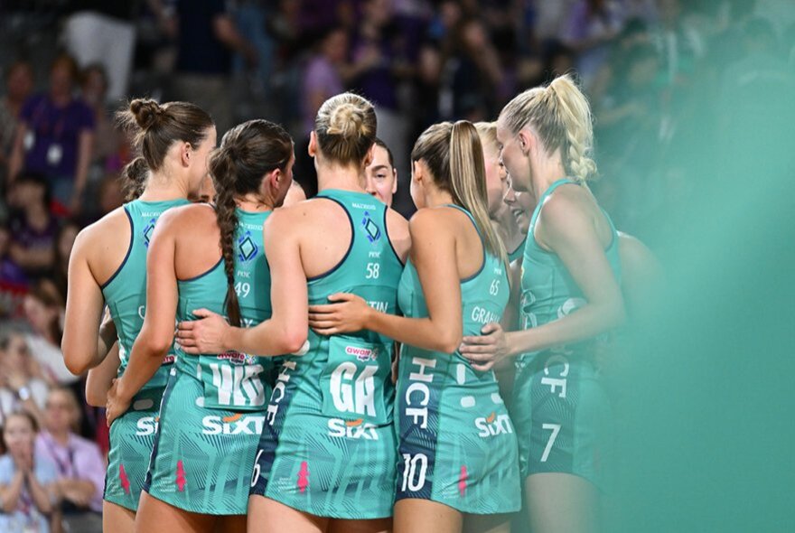 Women's netball team wearing teal uniforms, standing in a close circle during a game.
