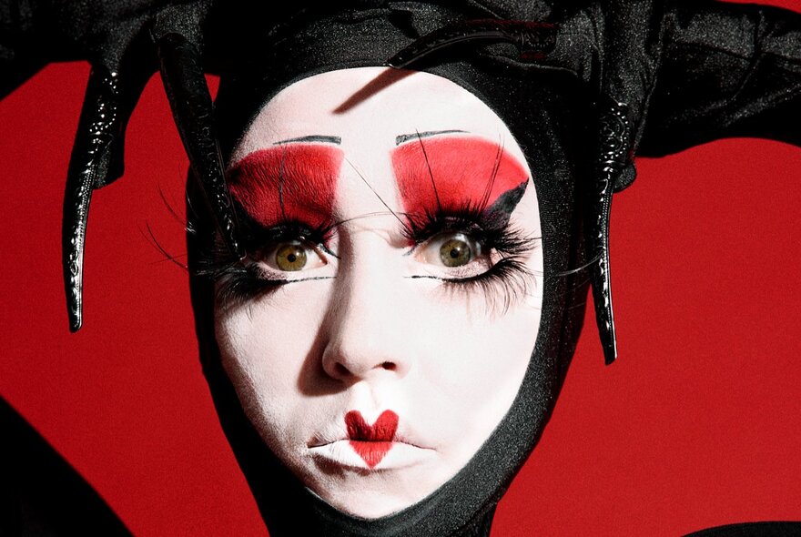 A woman's face heavily made up in white, red and black makeup, including a red heart on her lips and spidery black lashes.