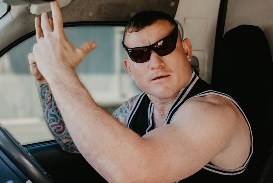 Comedian Mickey Wilson seated in a car with his arms up and looking gormless in a pair of speed-dealer sunglasses and a black singlet.