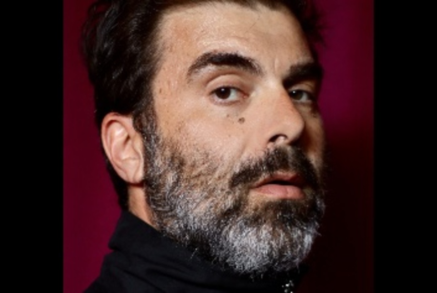 A close-up portrait of comedian, Evangelos Arabatzis, looking directly at the camera, with a full beard and wearing a black turtleneck top.