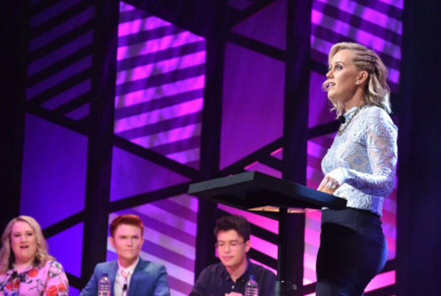 A comedian standing at a lectern talking, with the three members of the opposition debating team watching and listening.