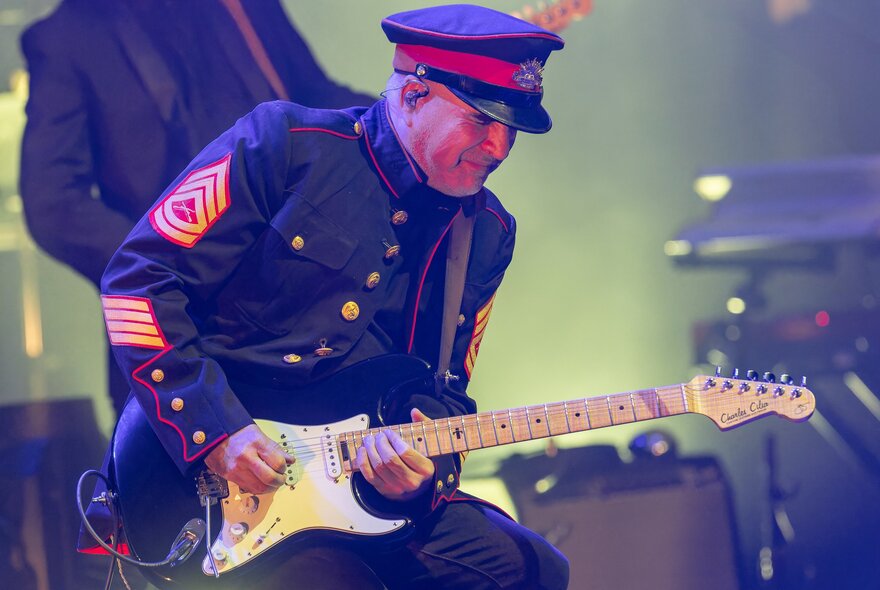 Guitarist performing live on stage wearing military-style coat and cap.