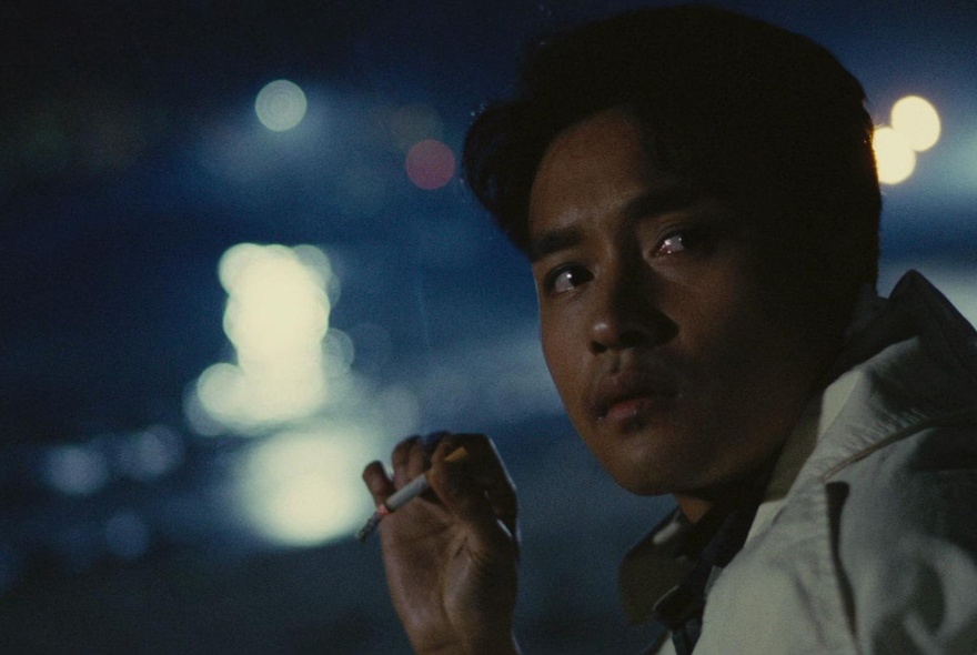 A night scene from the 1986 movie A Better Tomorrow, of a man in close-up profile, his head turned to face the viewer, holding a lit cigarette.