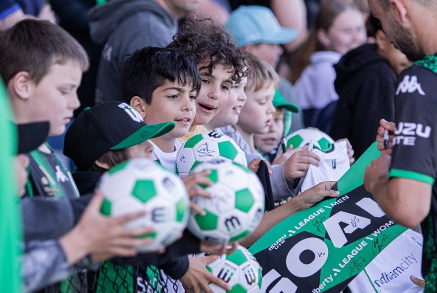 Young football fans in the stadium holding green and white soccer balls out to players for autograph signing. 