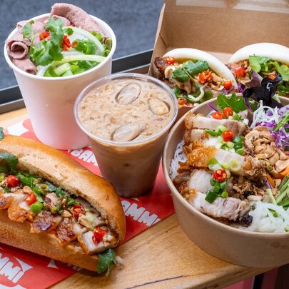 Looking down on a tray of Vietnamese lunch dishes including banh mi, vermicelli noodle bowl, bao buns, pho in a cup and Vietnamese iced coffee.