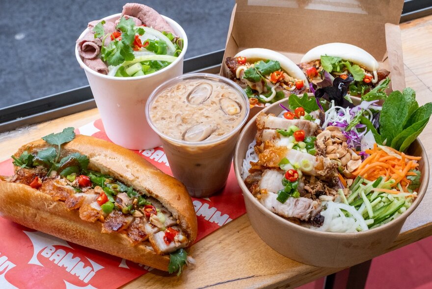 Looking down on a tray of Vietnamese lunch dishes including banh mi, vermicelli noodle bowl, bao buns, pho in a cup and Vietnamese iced coffee.