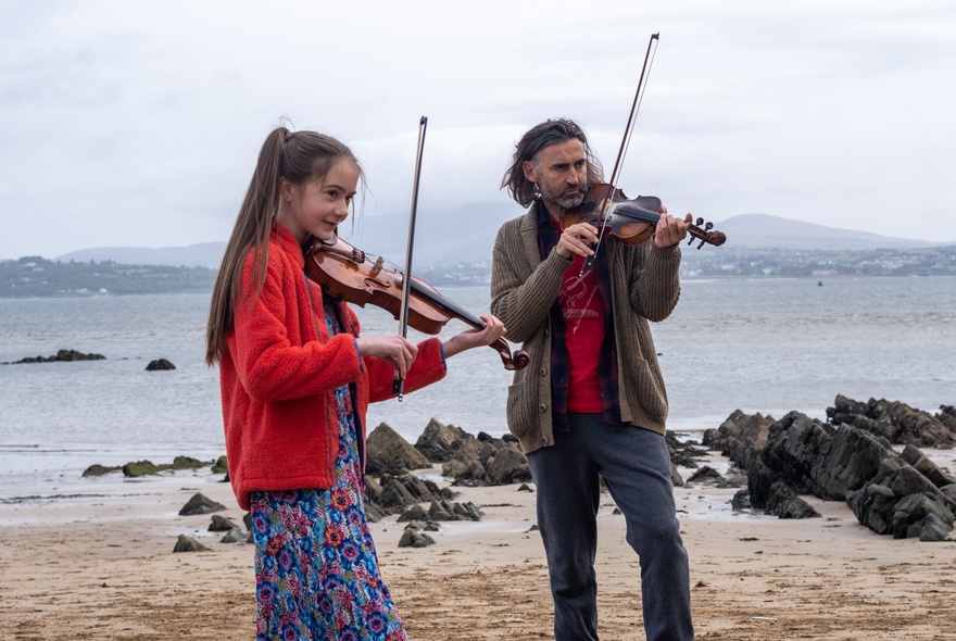A teenage girl and a young man both standing and playing fiddles on a beach, with a misty grey sea and horizon behind them.