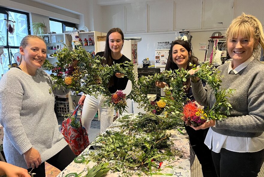 Several people smiling and standing in front of a work table covered in foliage clippings, holding up circular wreaths they've made using native flowers and greenery.