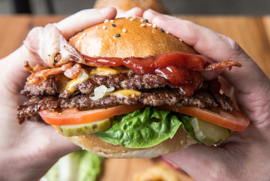 A double cheeseburger literally bursting with ingredients, being held by two hands.
