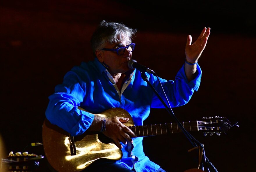 Italian musician, Fulvio Cama, wearing a blue suit and holding an acoustic guitar, on stage with a microphone.