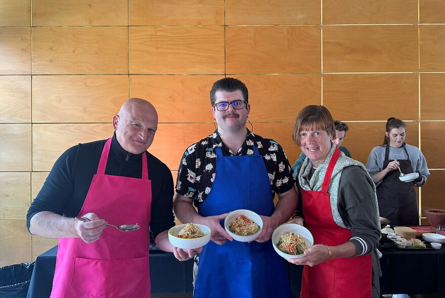 Three people wearing aprons, proudly showing a bowl of Thai food they prepared in  a cooking class.