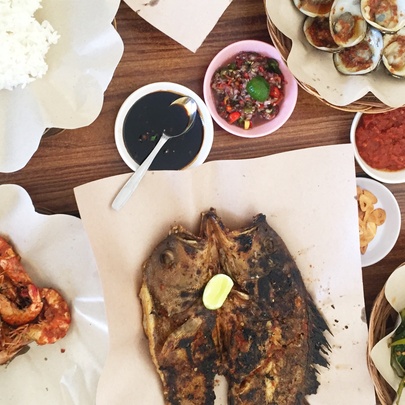 Looking down at a table of food, including grilled seafood, grilled prawns, and traditional Indonesian accompaniments like spicy sambal and stir-fried greens.