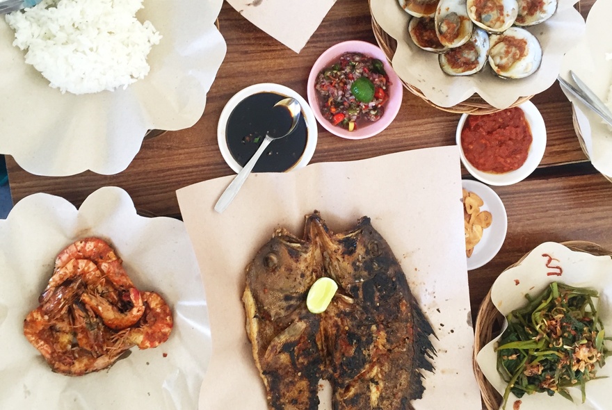 Looking down at a table of food, including grilled seafood, grilled prawns, and traditional Indonesian accompaniments like spicy sambal and stir-fried greens.