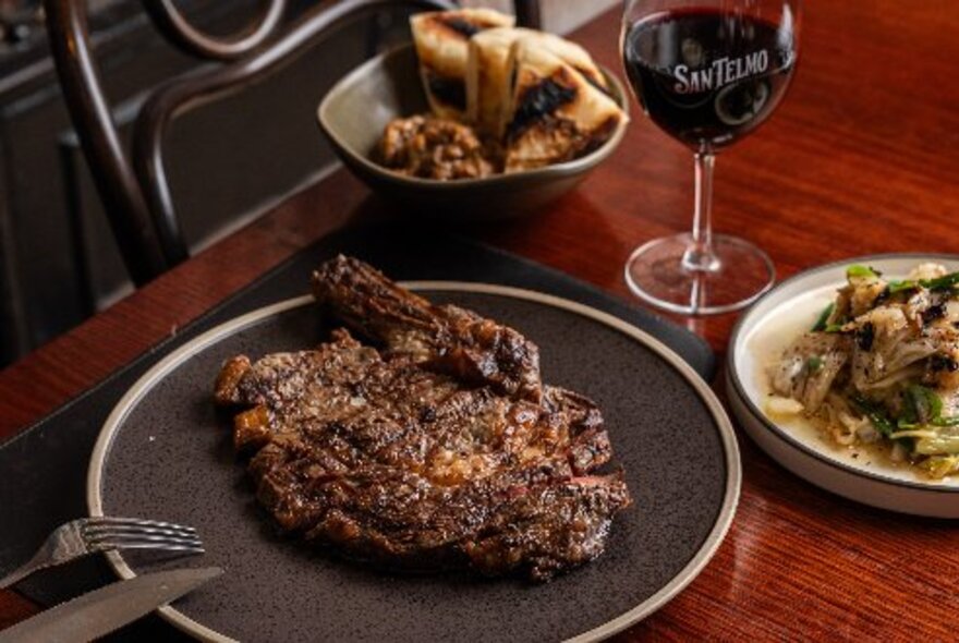 A large brown plate of cooked meat next to a glass of wine, bowl of bread and salad.