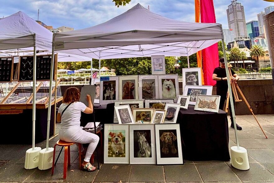 A stallholder at an outdoor market showcasing her framed artworks under a white gazebo.