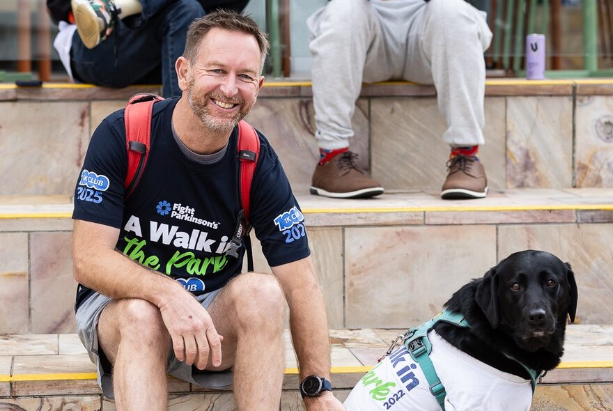 A smiling man sitting on tiled steps with his black labrador, both wearing t-shirts with the slogan, A Walk in the Park.
