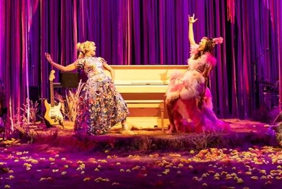 Two actors performing on a stage with purple curtains and a piano.