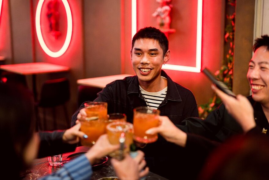 Smiling friends clinking glasses in celebration with neon and red decor. 