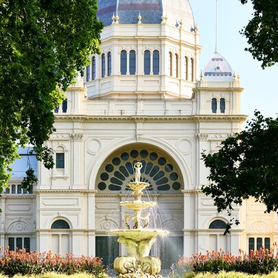 The grand arched entrance and central dome of the Royal Exhibition Building in Melbourne, viewed through a frame of green tree branches with a fountain in the centre foreground. 