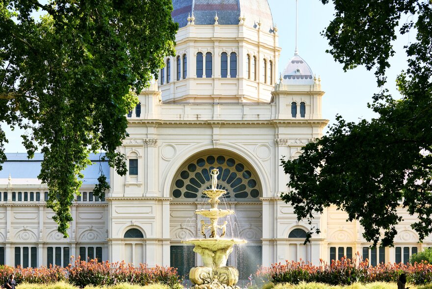 The grand arched entrance and central dome of the Royal Exhibition Building in Melbourne, viewed through a frame of green tree branches with a fountain in the centre foreground. 