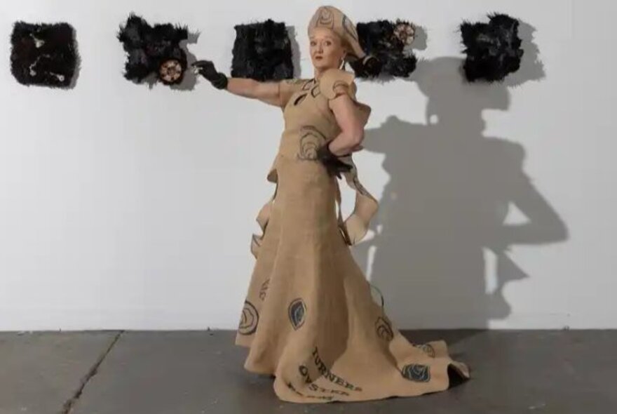 Woman wearing ragged beige gown, standing in front of artwork installation on wall.