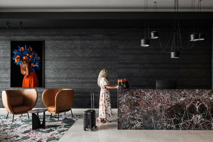 Person with suitcase standing at a hotel foyer with large granite desk, lounge seating, pendant lighting and feature painting.