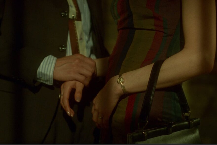 Two people standing next to each other, their faces out of view, the man's hand gently holding the wrist of the woman, who carries a handbag over her other forearm.