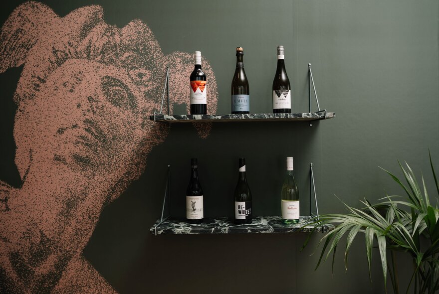 Two floating shelves mounted on a dark green wall, each holding three bottles of wine, with a stylized portrait of a face painted in a reddish-brown on the wall.